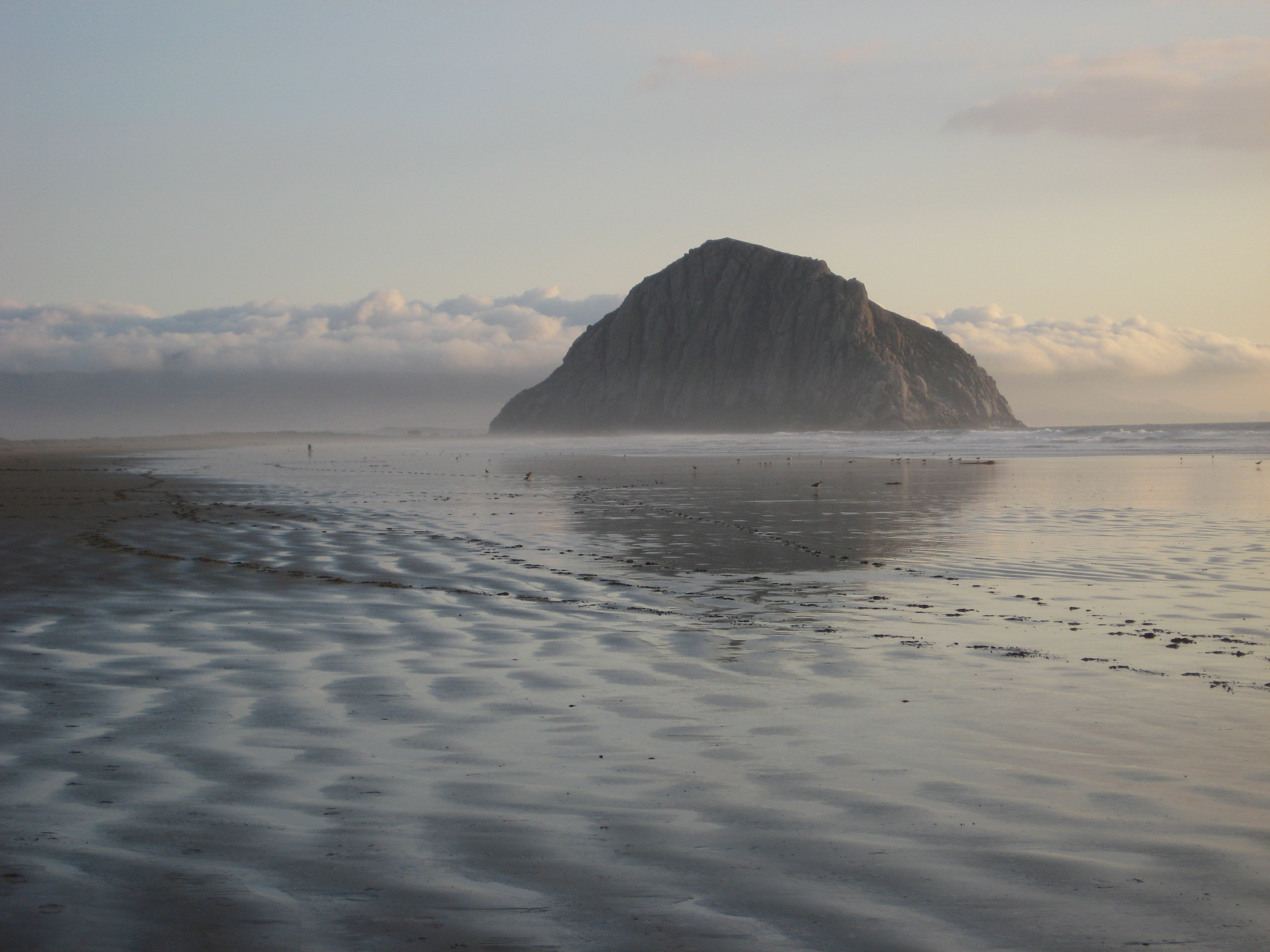 Morro Rock reflected in rippled wet sand and backed by fluffy white clouds on a dreamy evening.