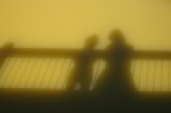 Shadows of two people on a bridge reflect in murky green water.