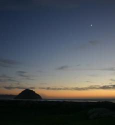 A crescent moon hangs in the night sky above Morro Rock. 