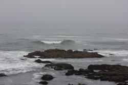 A small wave crests near the rocky shore, against a foggy blurred horizon.